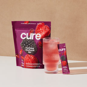 Cure Hydration Electrolyte Mix package with images of berries and watermelon, next to a glass of pink beverage and a single serving stick pack