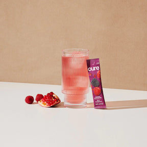 Glass of pink lemonade with a Cure berry pomegranate drink mix packet and fresh raspberries and pomegranate seeds beside it