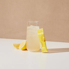 Glass of lemonade with ice, lemon slices, and a yellow 'cure' packet on a white surface with a beige background