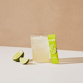 Glass of lime drink with ice, lime wedges, and a green packet labeled 'cure' featuring lime illustrations