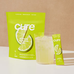 Package of Cure Hydrating Electrolyte Mix in Lemon Lime flavor, with a glass of the prepared drink and a single stick pack beside it