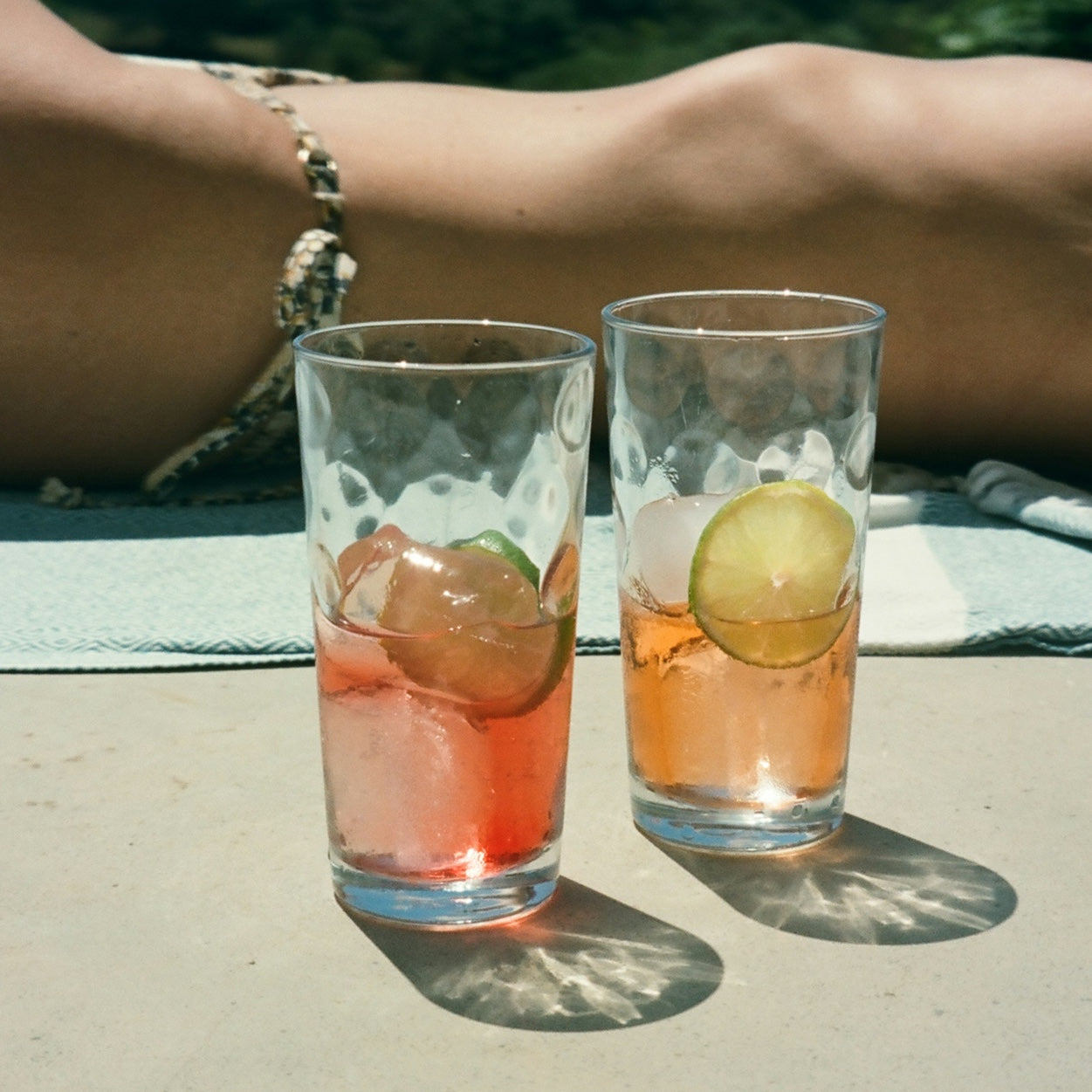 Two glasses with ice, lime slices, and pinkish electrolyte drinks on a table, with a person's legs in the background.