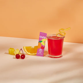 A glass of red juice with a yellow curly straw, a pink box labeled 'cure kids variety pack', a slice of orange, two cherries, and two pineapple chunks on a beige surface.