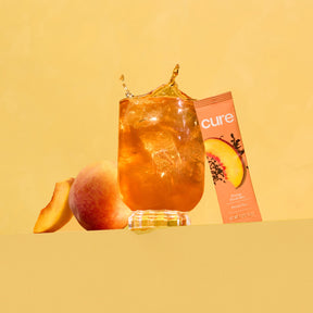 Glass of iced peach drink with a peach fruit, a peach slice, and a peach-flavored product box labeled 'cure'