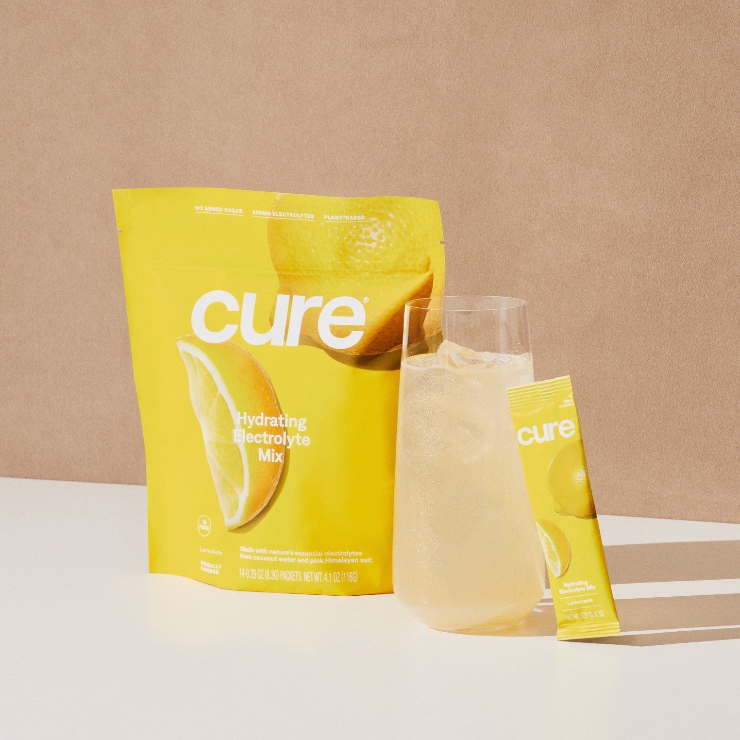 Yellow packet labeled 'cure hydrating electrolyte mix' with lemon graphics, a glass of lemonade, and a single stick packet labeled 'cure'.