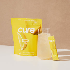 Yellow packet labeled 'cure hydrating electrolyte mix' with lemon graphics, a glass of lemonade, and a single stick packet labeled 'cure'.