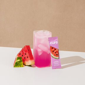Pink iced drink next to watermelon slices and a purple packet labeled 'cure' with watermelon image