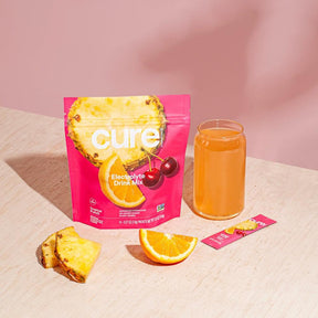 Cure brand Tropical Punch/Berry Pomegranate Bundle packaging with pineapple slice, cherries, a glass of orange drink, pineapple and orange fruit slices, and a single stick pack on a light surface with pink background